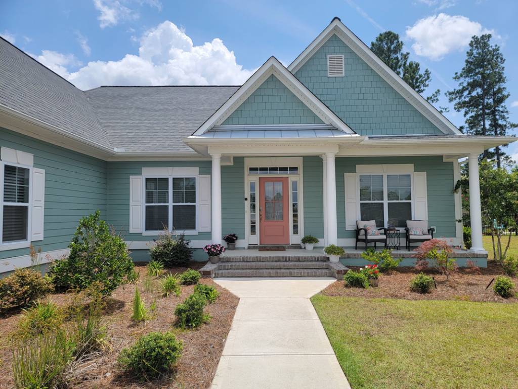Light green coastal home with coral front door, white shutters, and manicured landscaping on a sunny day.