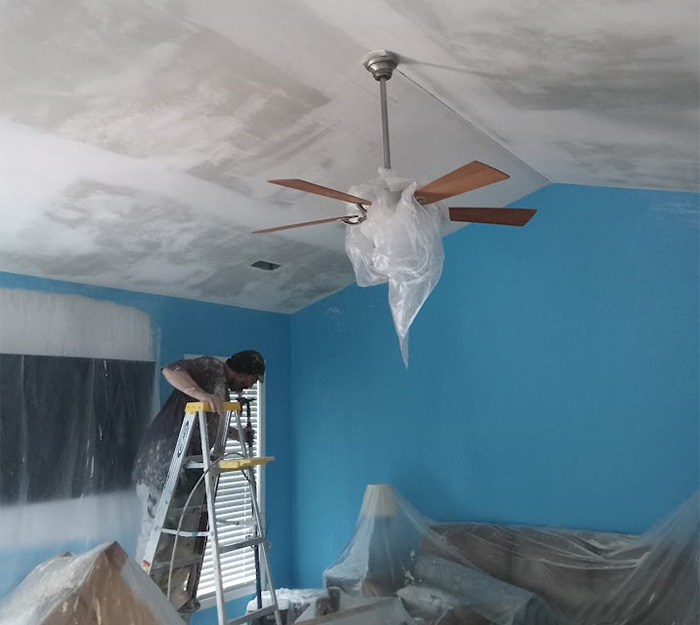 Worker repairing and painting interior ceiling with protective plastic covering in a blue room – JBG Painting.