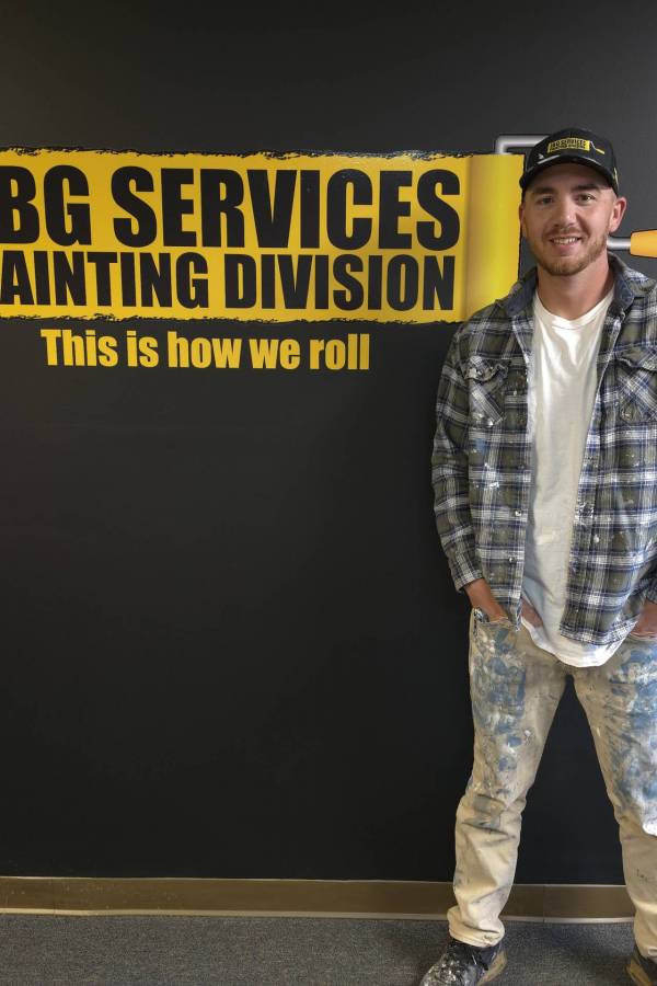Robert, a JBG Painting team member, smiles in front of the JBG Services Painting Division branded wall, wearing a JBG cap, plaid shirt, and paint-covered work pants.