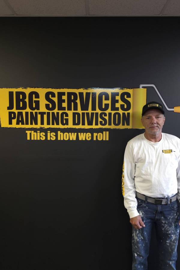 Sean, a JBG Painting team member, stands in front of the JBG Services Painting Division branded wall, wearing a JBG cap, company long-sleeve shirt, and paint-splattered jeans.