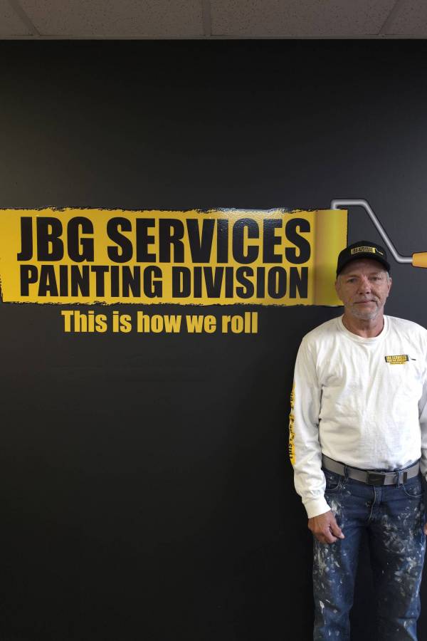 Sean, a JBG Painting team member, stands in front of the JBG Services Painting Division branded wall, wearing a JBG cap, company long-sleeve shirt, and paint-splattered jeans.