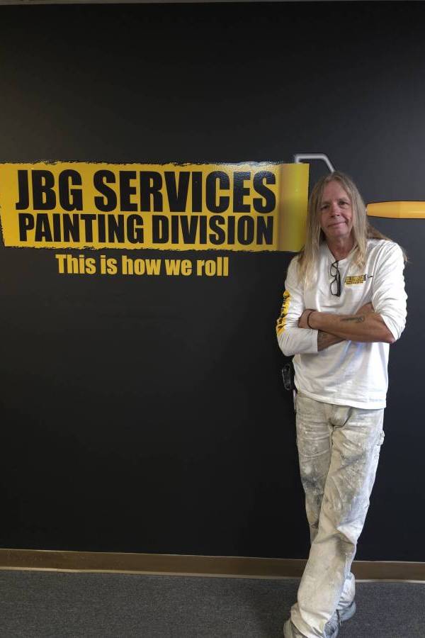 Will, a JBG Painting team member, stands in front of the JBG Services Painting Division branded wall in Wilmington, NC, wearing a JBG uniform shirt and paint-spattered work pants.